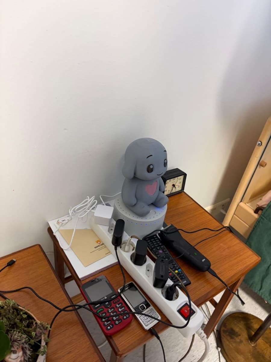 A grey Qluve robot on a resident's bedside table, next to an alarm clock and two mobile phones